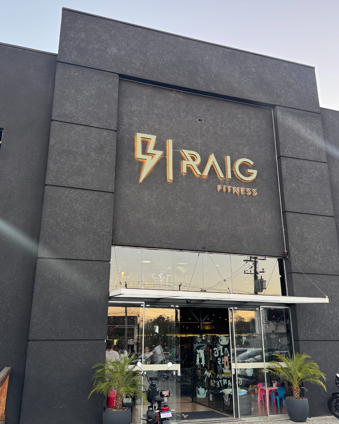 Fachada da Raig Fitness na Av. Brasília, no Capão Raso, com o letreiro iluminado do raio ao anoitecer